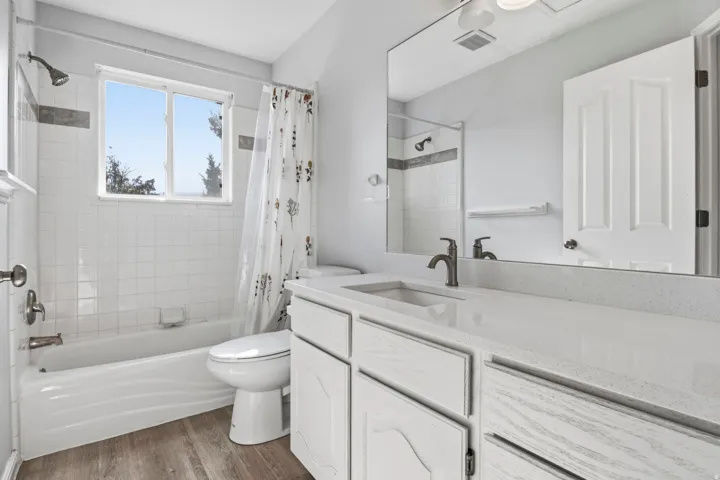 White tile surround tub and shower with a window, single vanity with an integrated sink, large mirror, and wood-finish flooring