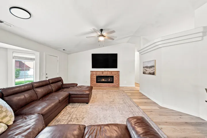 Vaulted ceilings with a ceiling fan, brick fireplace surround with an electric insert, wood-finish flooring, and white interior walls