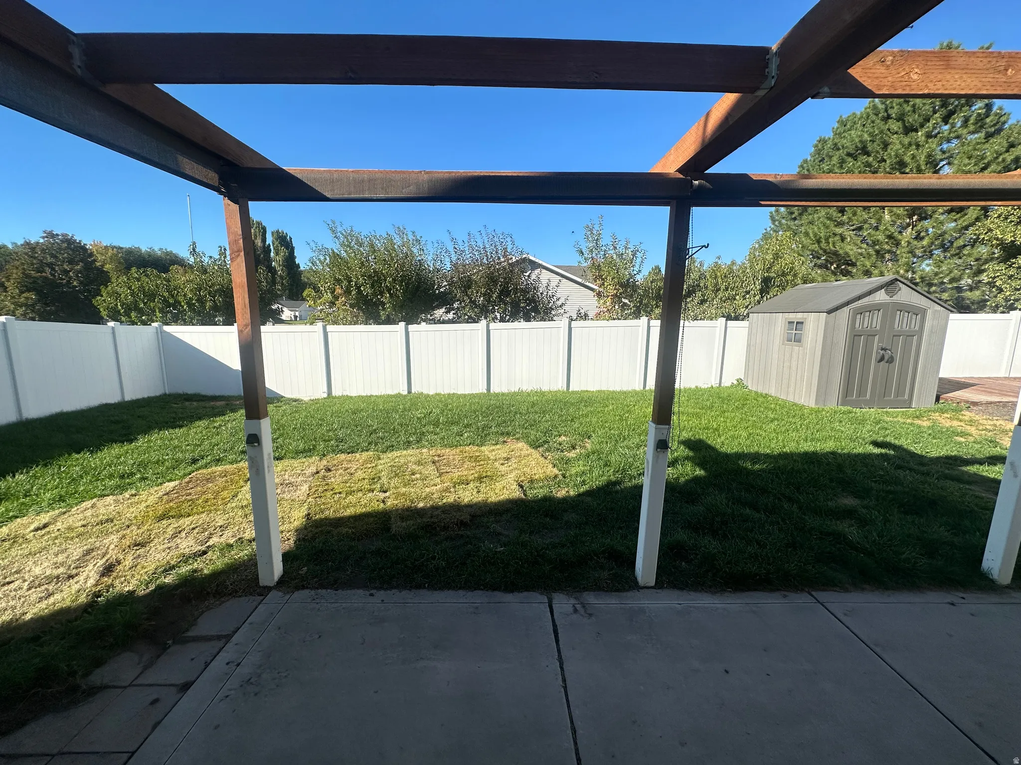 Patio area with concrete surfacing, wood pergola structure, green lawn, white privacy fencing, and a storage shed