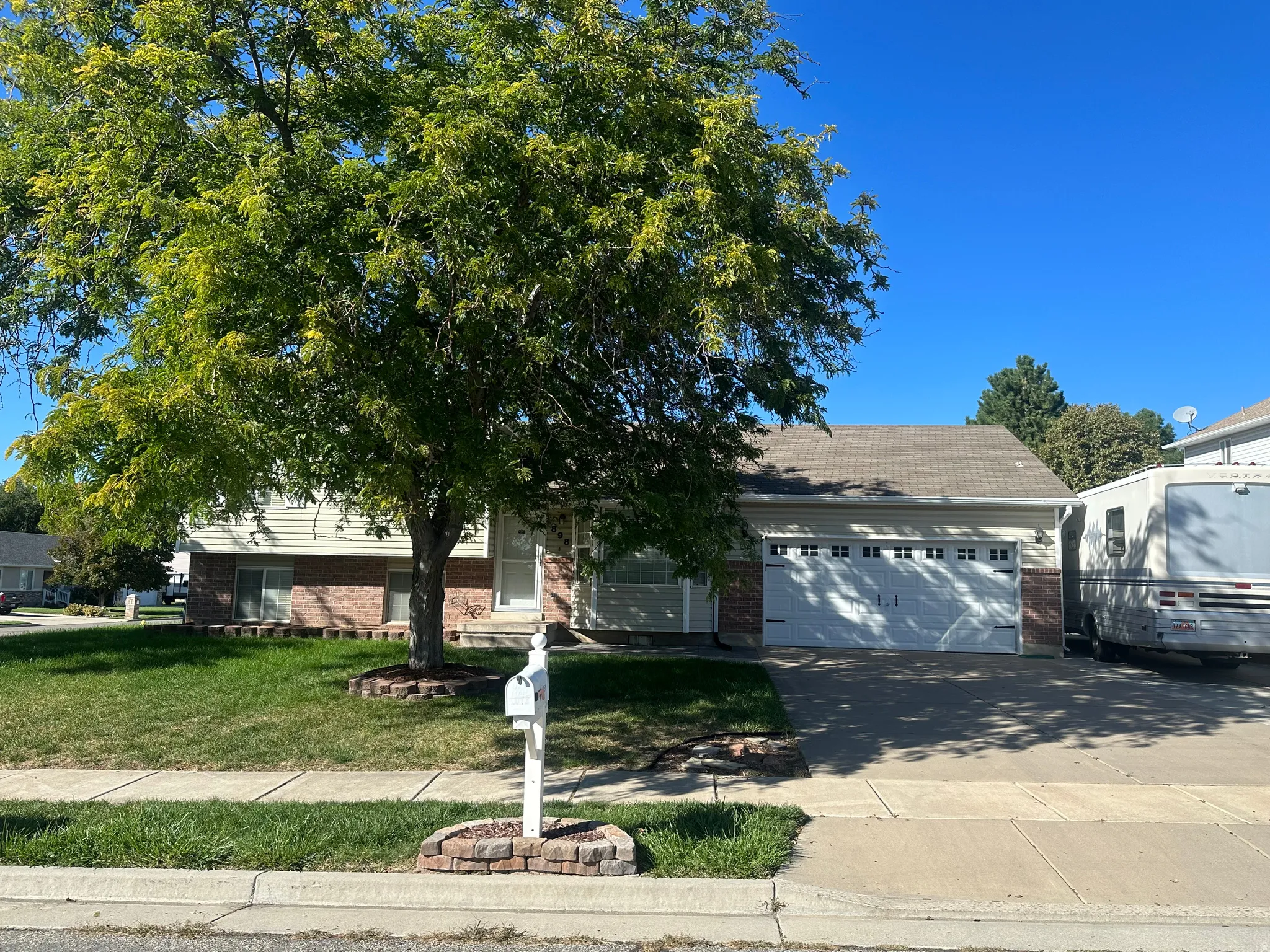Manicured front yard with mature tree, brick-accented exterior, two-car garage, and concrete driveway