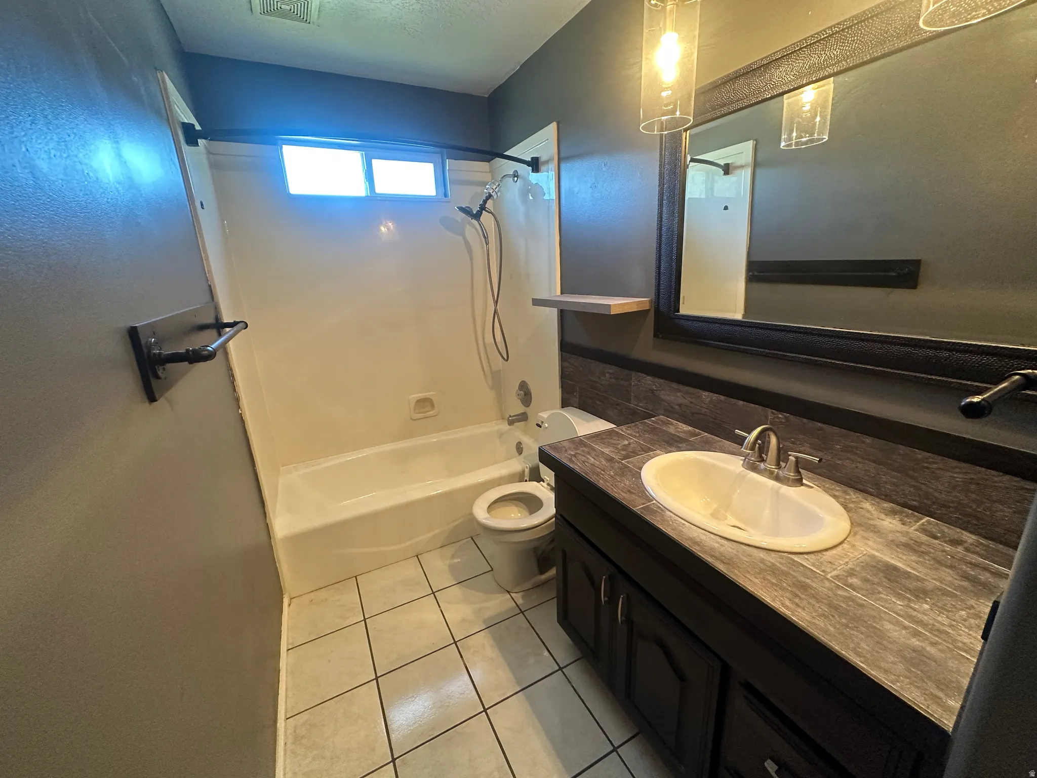 Tiled flooring, white bathtub, dark wood-finish vanity with integrated sink, framed mirror, and pendant lighting fixture