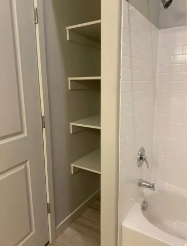 Built-in shelving, white subway tile shower surround, white bathtub, chrome shower fixture, wood-finish flooring