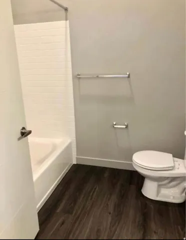 Tub and shower combination with white subway tile surround, wood-finish flooring, and a white toilet