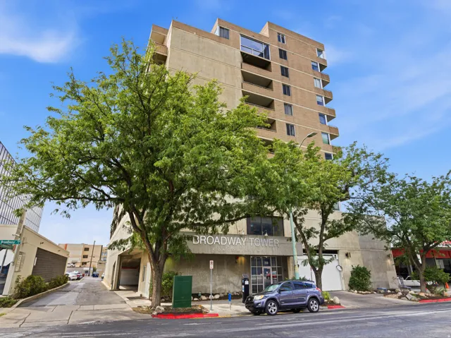 Mid-rise residential building featuring a concrete and masonry facade with multiple balconies, ground-level retail space, and mature landscaping including established trees flanking the street frontage