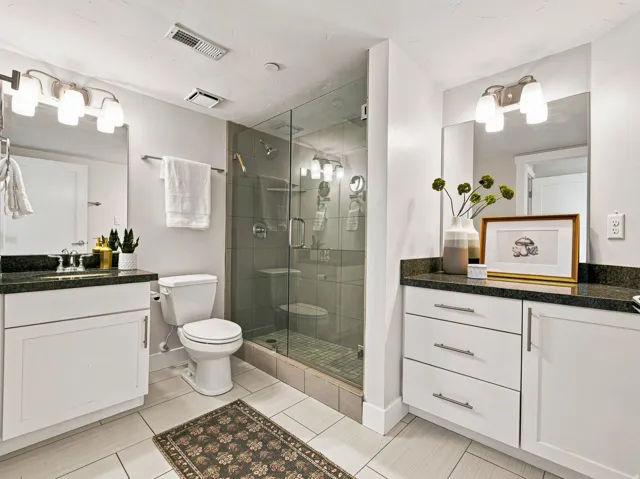 Dual vanities with dark countertops, a glass-enclosed shower, white cabinetry, and light-toned tile flooring