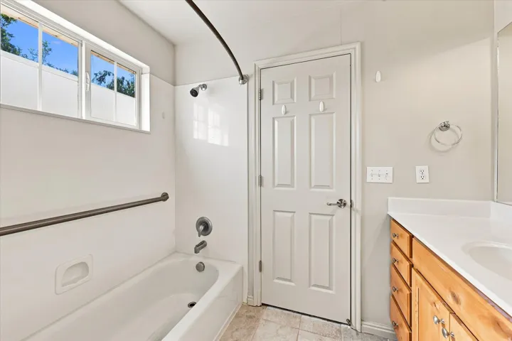 White bathtub with shower, horizontal window, wood-finish vanity with white countertop, and neutral wall paint