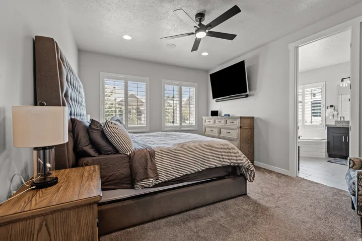 Carpeted bedroom featuring two windows with white plantation shutters, a ceiling fan with integrated lighting, and recessed ceiling lights