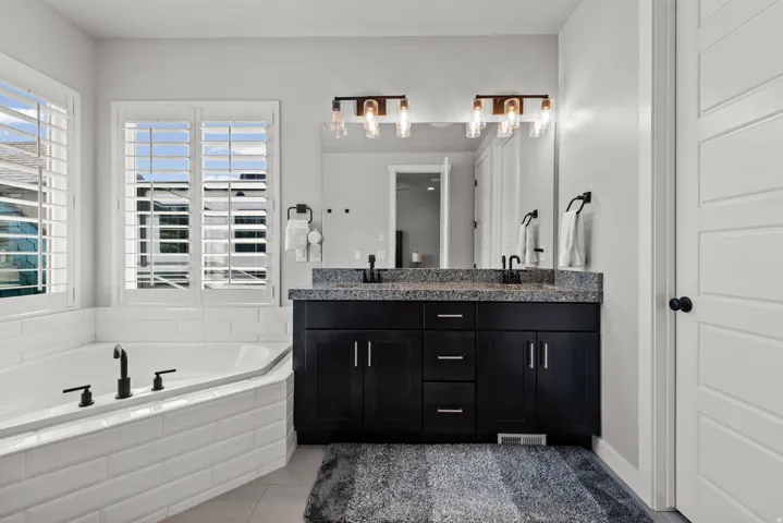 Soaking tub with white subway tile surround, dual window with plantation shutters, black double vanity with stone countertop, and two sets of bronze-finish light fixtures