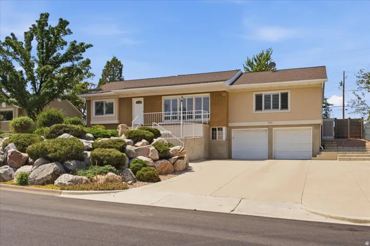 Split-level home featuring a stucco exterior with a covered front porch, dual two-car garage with concrete driveway, and professionally landscaped front grounds with mature trees, decorative boulders, and manicured shrubs