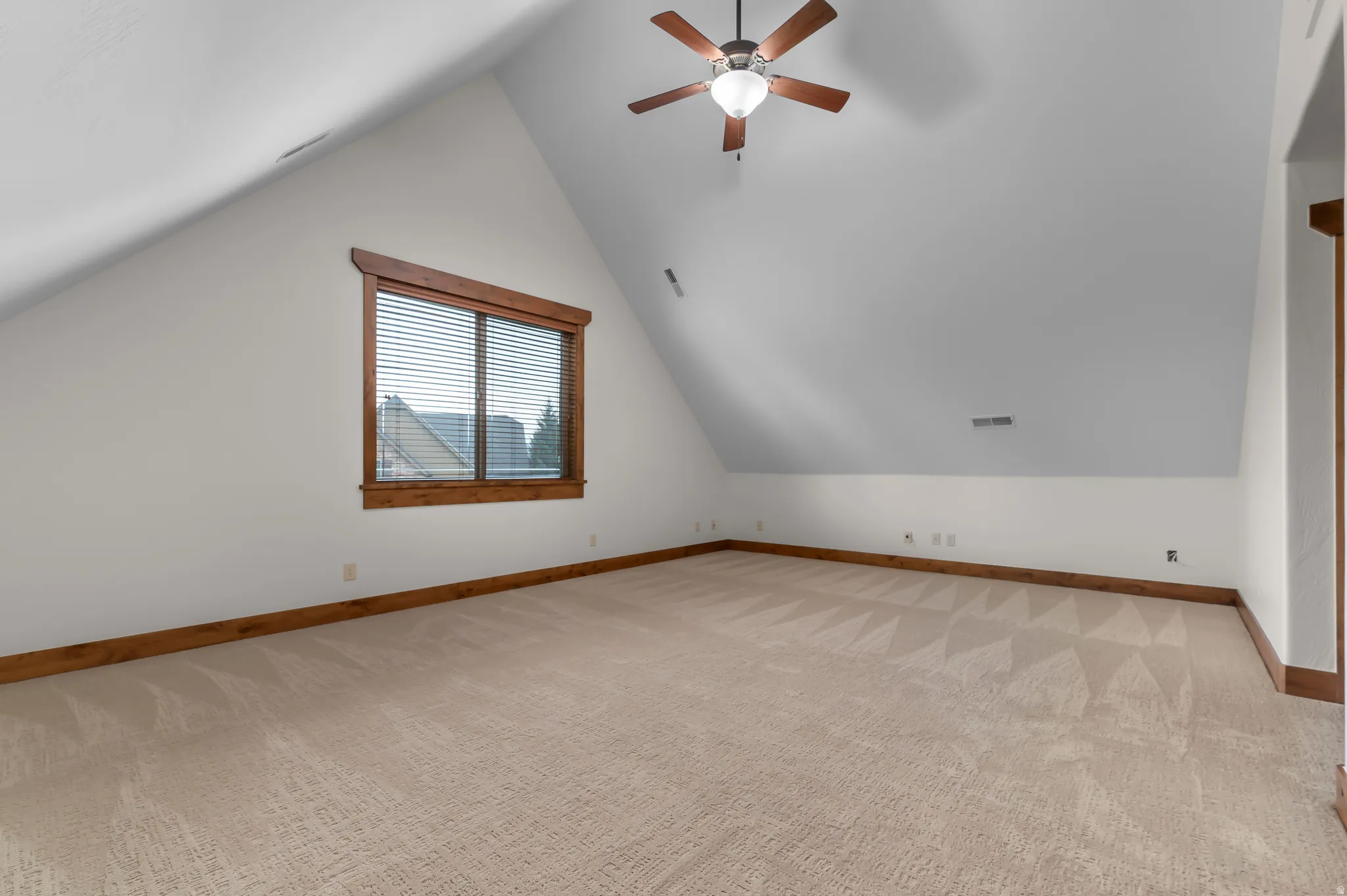 Spacious room with sloped ceilings, a ceiling fan, a window with wood trim, and light-toned carpeting