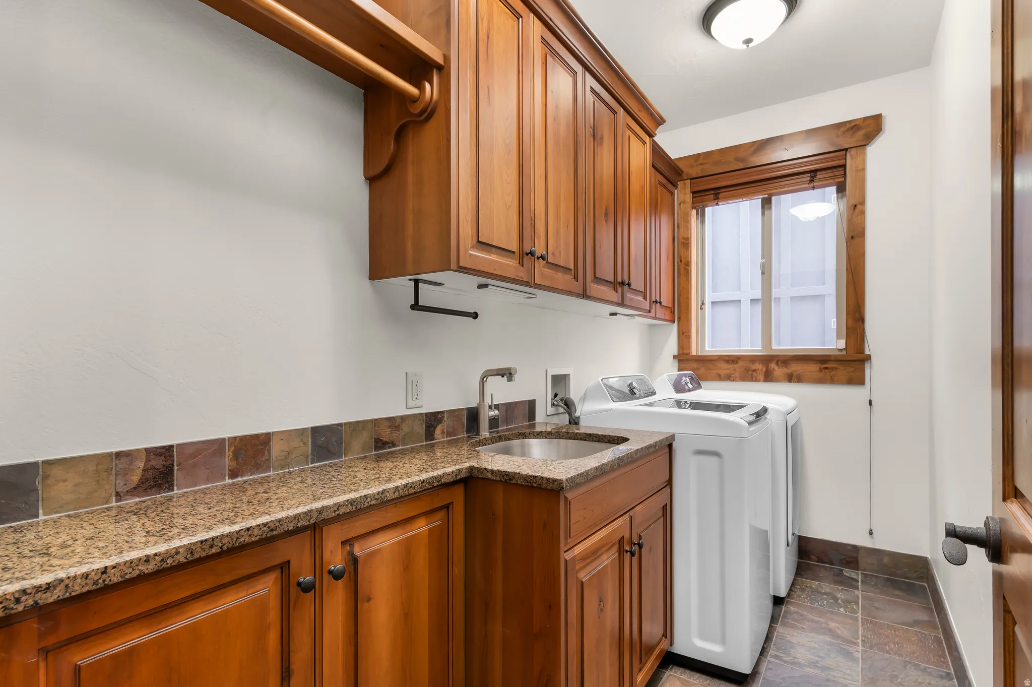 Laundry area featuring wood cabinetry, stone-finish countertops, an undermount sink, and a tiled backsplash