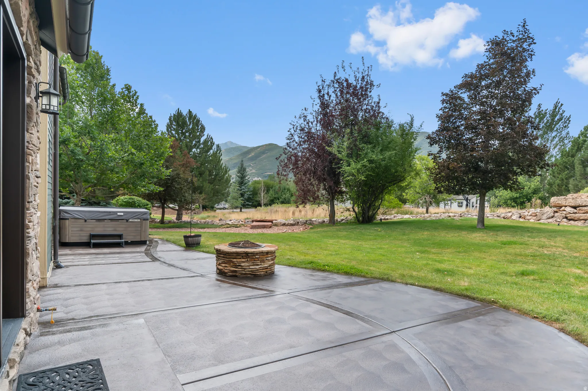 Expansive backyard featuring a large concrete patio, stone fire pit, and mature trees, with mountain scenery