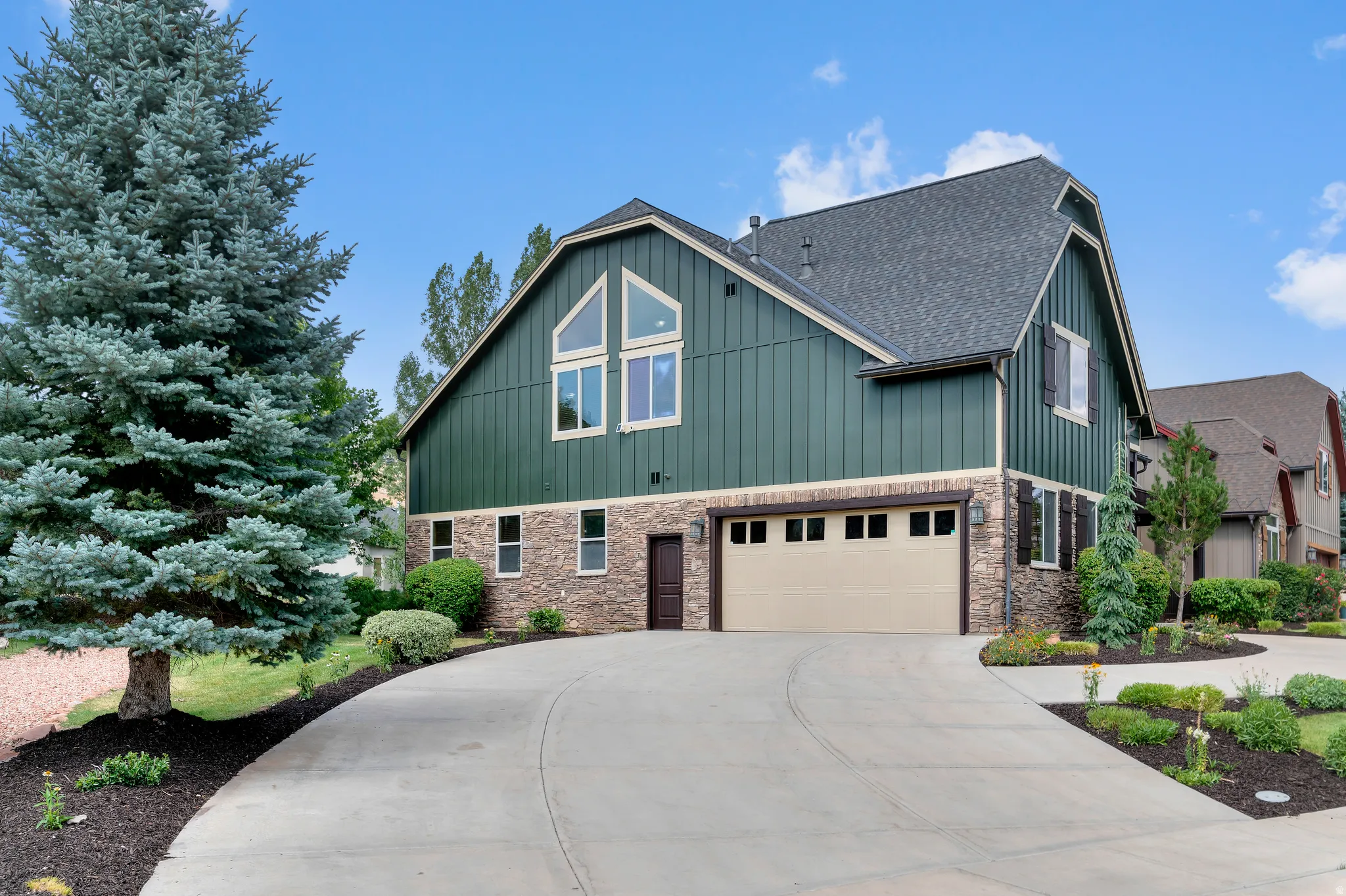 Exterior featuring green vertical siding, stone accents, multiple large windows, a two-car garage, and a concrete driveway