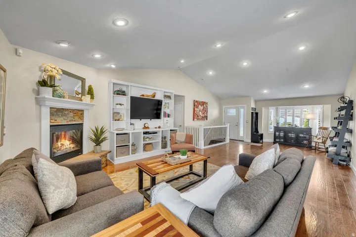 Vaulted ceiling with recessed lighting, wood-finish flooring, stone-surround fireplace with a white mantel, built-in media console, and a bay window