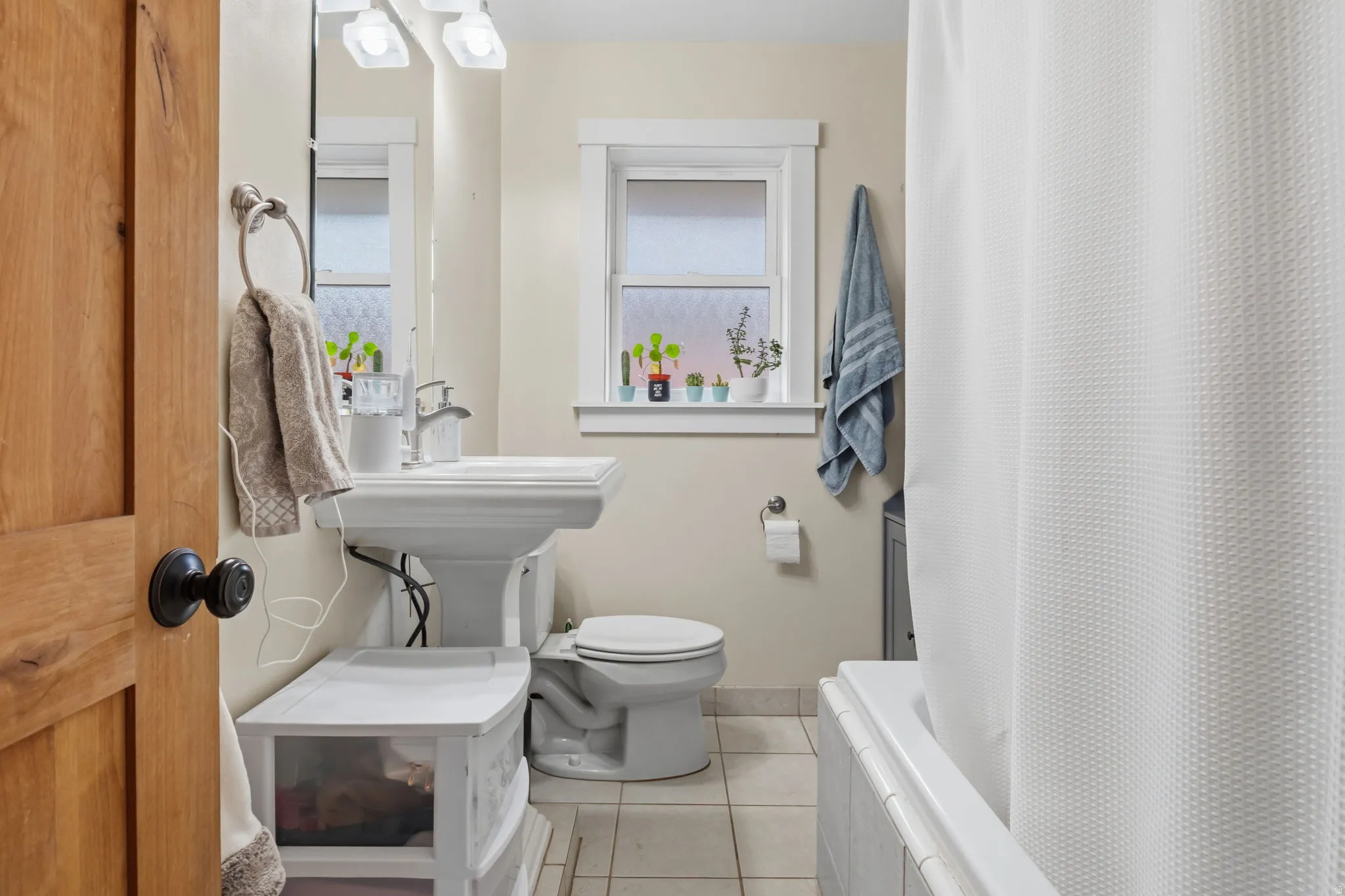 Pedestal sink with chrome faucet, tiled flooring, window with frosted glass, and white bathtub with tiled surround