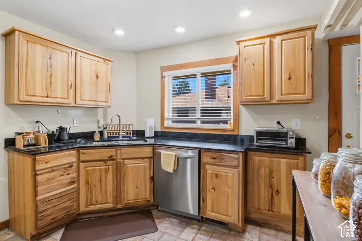 Kitchen featuring wood cabinetry, dark countertops, a stainless steel dishwasher, and a window with blinds