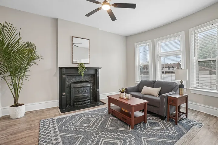 Living area with wood-finish flooring, a decorative fireplace, multiple windows with blinds, and a ceiling fan with integrated lighting