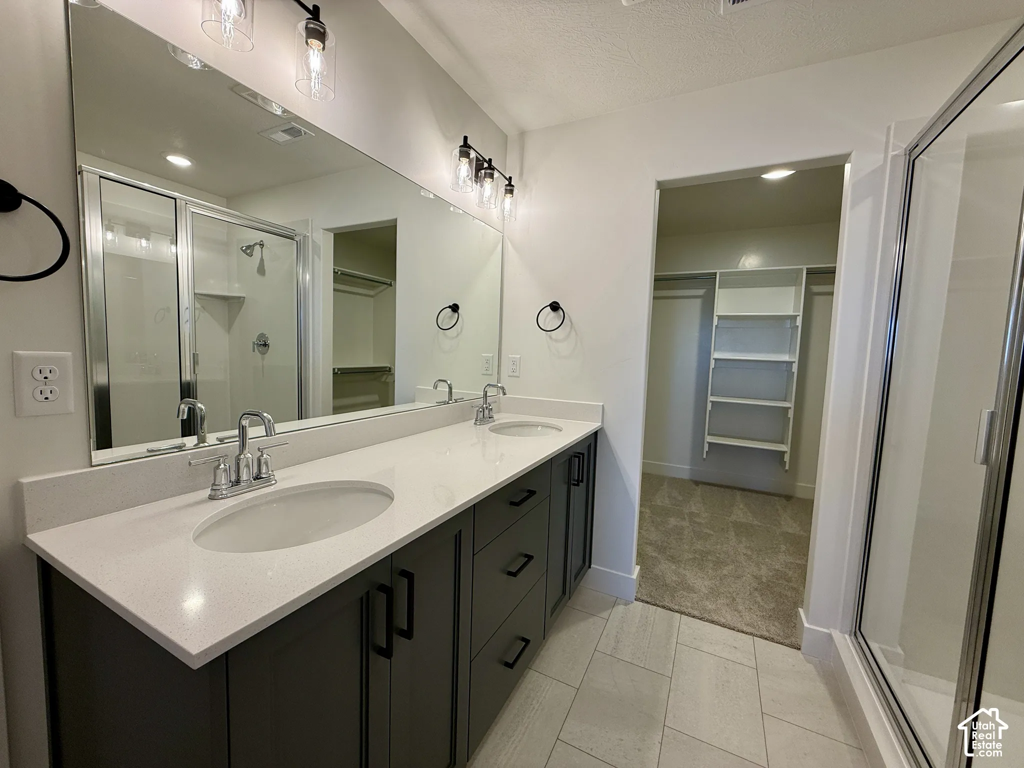 Dual-sink vanity with a quartz countertop, dark-finish cabinetry, full-width mirror, glass-enclosed shower, and built-in closet shelving