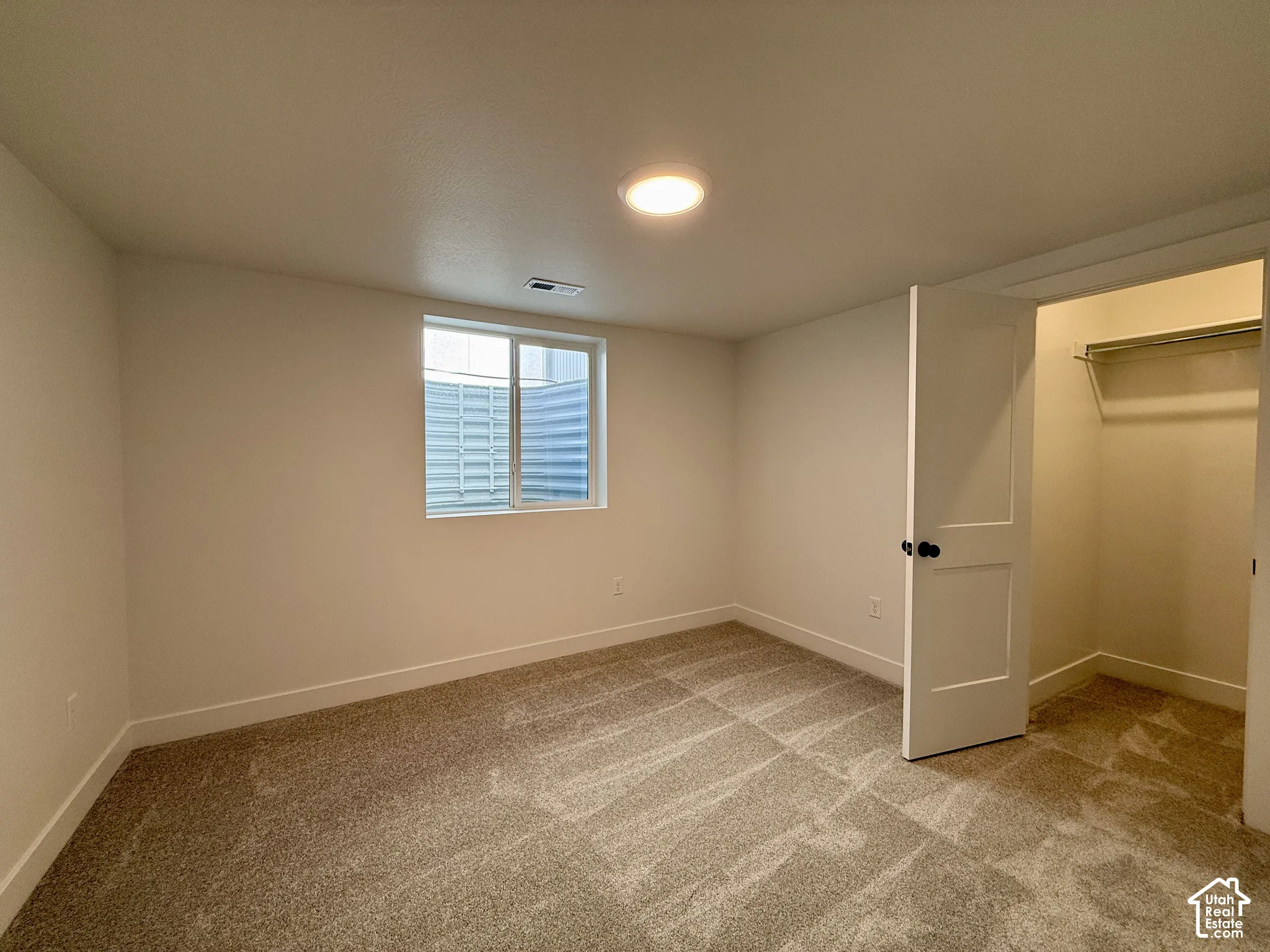 Neutral-toned carpeting, flush-mount ceiling lighting, sliding egress window with metal window well, and interior closet featuring a built-in shelving rod