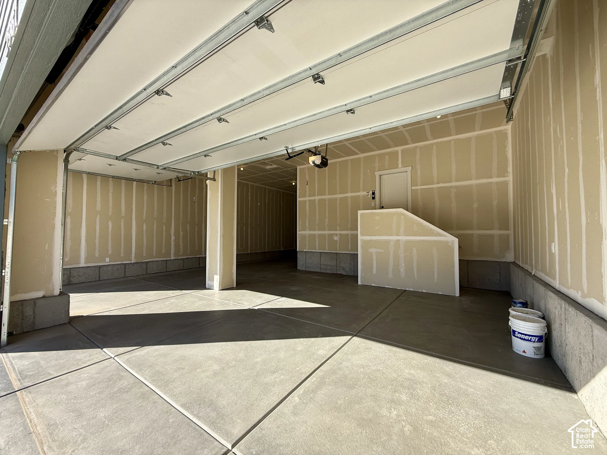 Spacious multi-car garage featuring a concrete floor, overhead door mechanism, and drywall-finished walls with a built-in partition structure