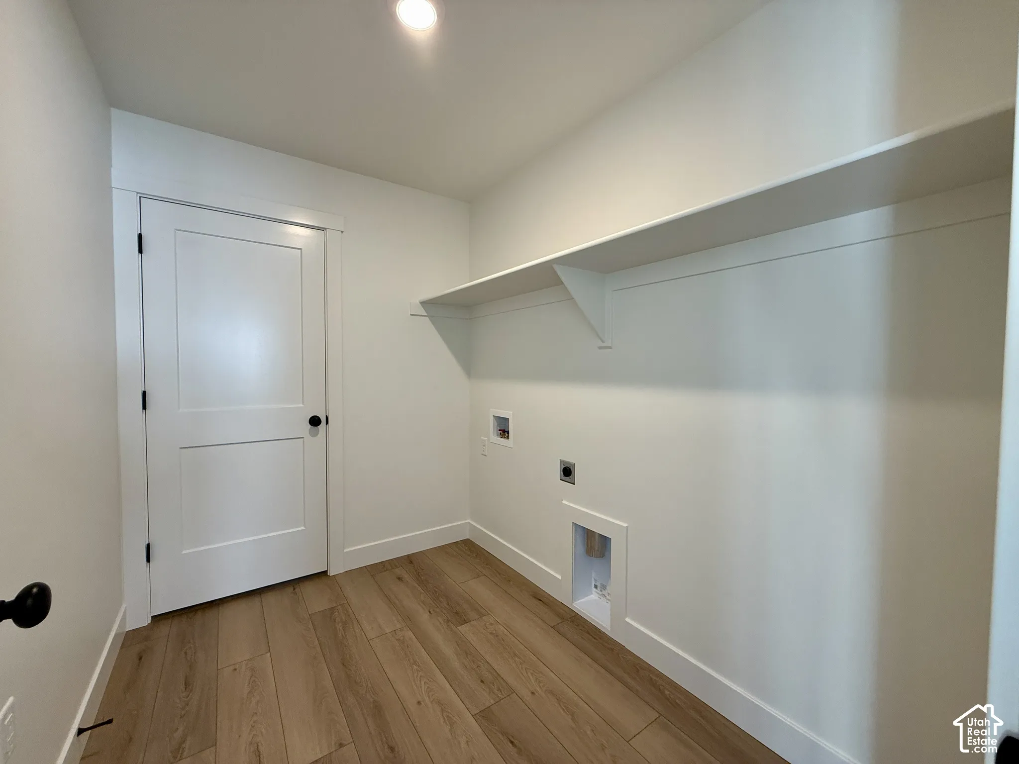 Utility space featuring wood-finish flooring, a white two-panel interior door with matte black hardware, recessed ceiling lighting, and a full-width wall-mounted shelf