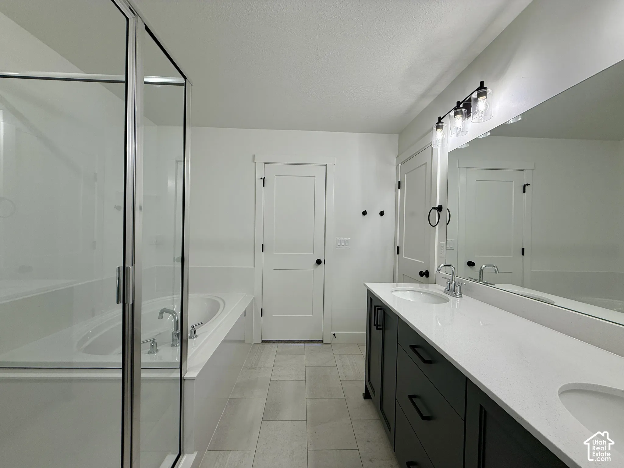 Double-sink vanity with dark cabinetry, quartz countertops, soaking tub, glass-enclosed shower, and large-format tile flooring