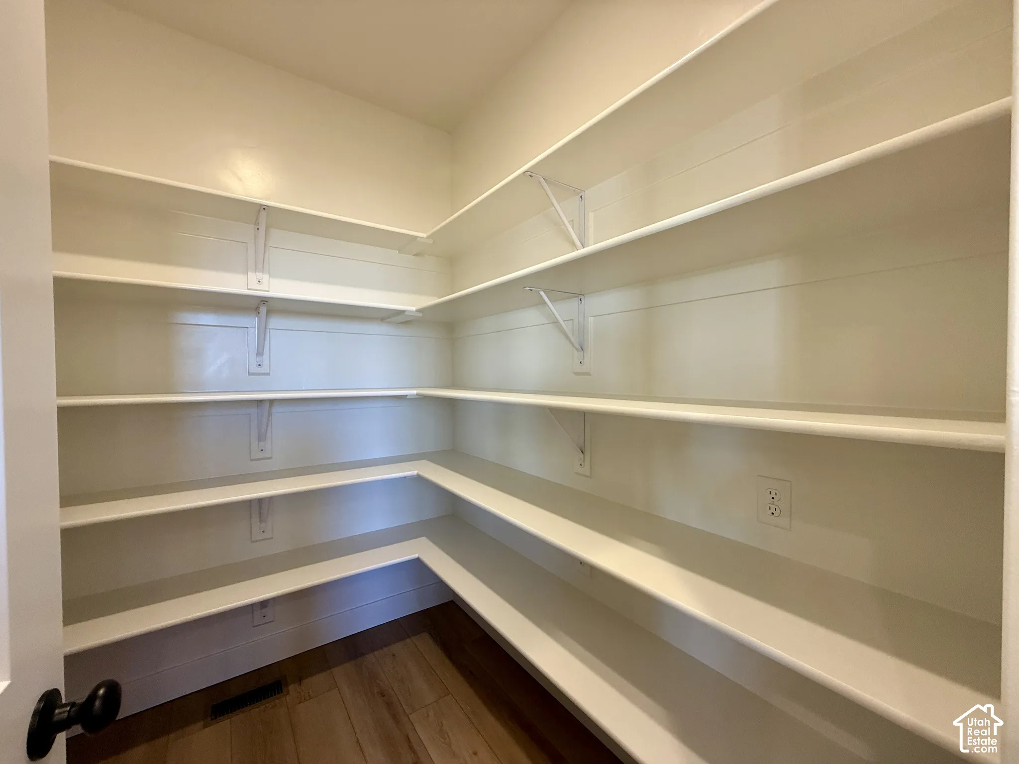 Walk-in storage space featuring multi-tiered white shelving, wall-mounted supports, and wood-finish flooring