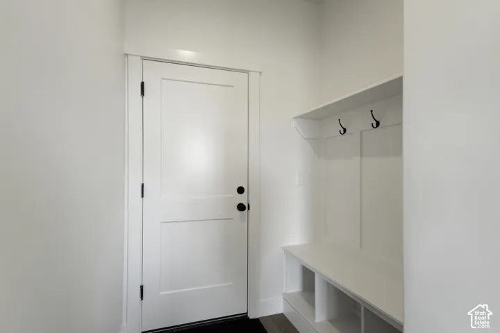 Bright entryway featuring a clean-lined white six-panel entry door with oil-rubbed bronze hardware, light-filled walls, and a custom built-in mudroom with white cabinetry, open shelving, and dual wall-mounted hooks for convenient storage and organization