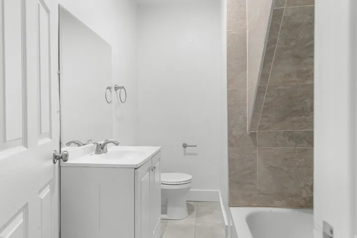 White vanity with an integrated sink and chrome faucet, large frameless mirror, and a tiled shower-tub combination