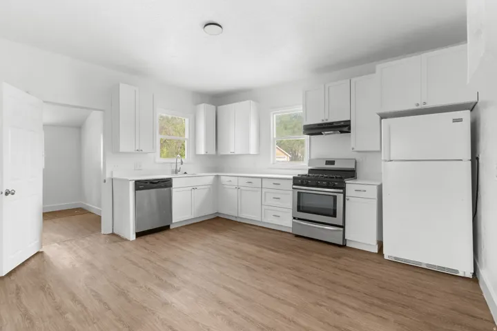 Bright kitchen featuring white cabinetry, stainless steel appliances including a double wall oven and gas cooktop, white refrigerator, wood-finish flooring throughout, and abundant natural light from multiple windows