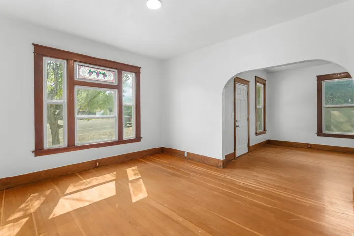 Bright, open living space featuring hardwood flooring throughout, expansive windows with wood-frame trim flooding the room with natural light, and an arched architectural detail connecting to an adjacent room