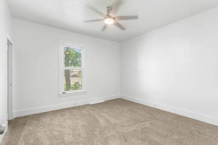 Bright and spacious bedroom with neutral carpet flooring, large window with natural light, ceiling fan with integrated lighting, and clean white walls throughout