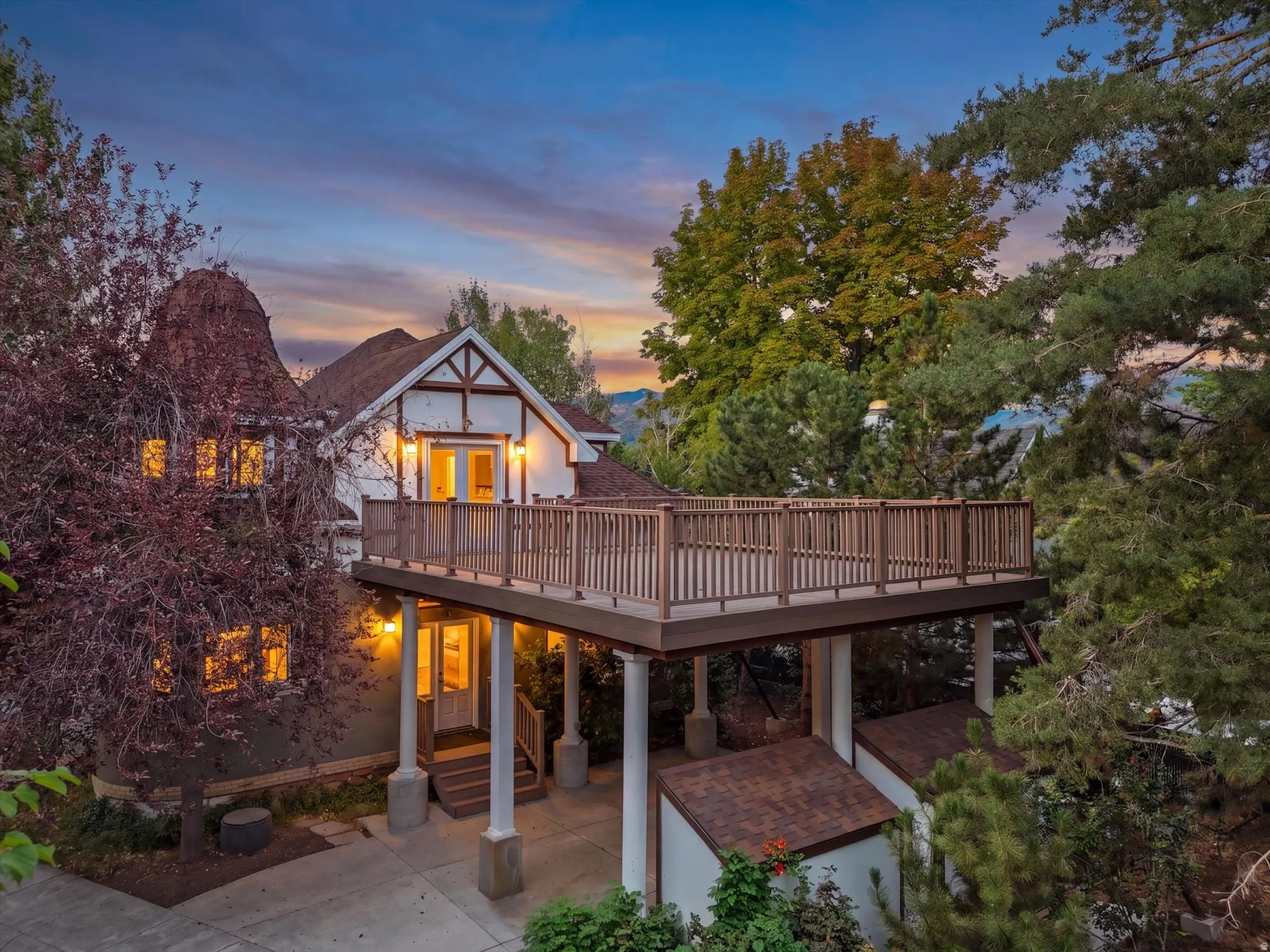 Charming Tudor-style residence with illuminated interior spaces, expansive composite deck with white railings, and mature landscaping throughout the grounds