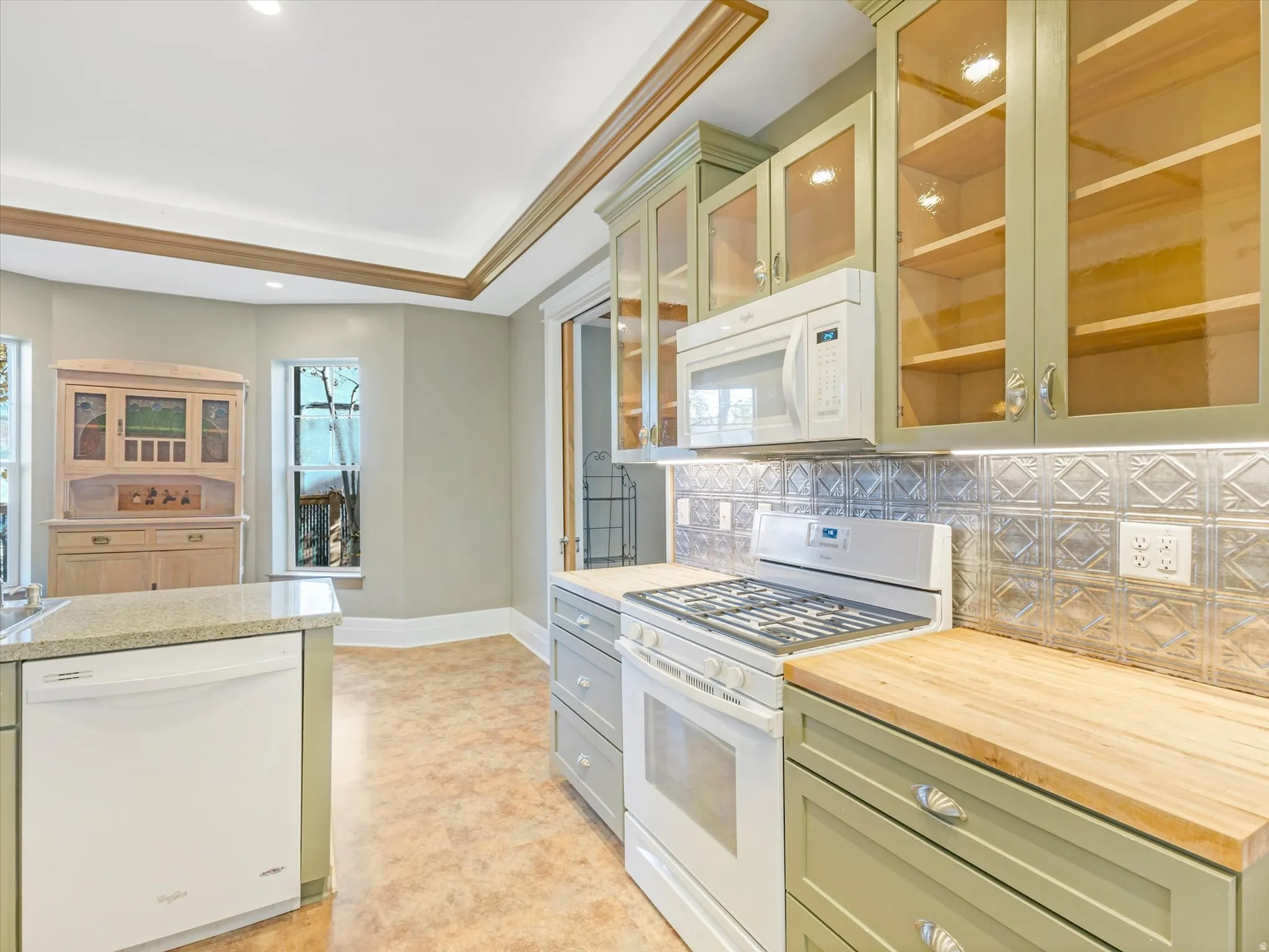 Kitchen featuring light green cabinetry with glass-front upper doors, wood-finish countertops, stainless steel appliances, and a textured metal backsplash