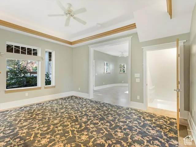 Recessed ceiling with wood trim, ceiling fan, multiple windows, patterned carpet, and an open doorway