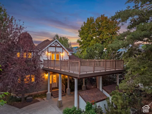 Charming Tudor-style residence with illuminated interior spaces, expansive composite deck with white railings, and mature landscaping throughout the grounds