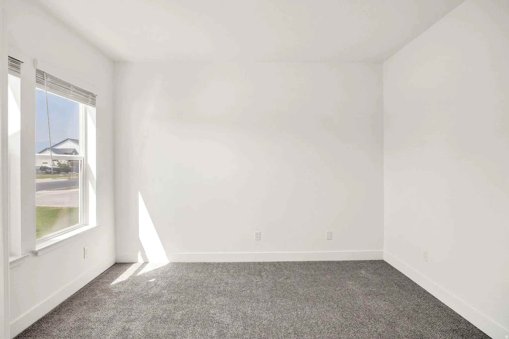 White painted walls with gray carpet flooring, featuring a large window with blinds and white trim