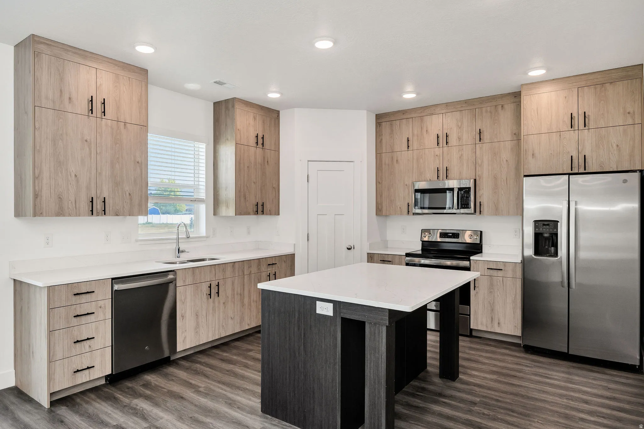 Modern kitchen featuring custom wood-finish cabinetry with brushed metal hardware, white quartz countertops, a large center island with black wood-finish base, stainless steel appliances including a refrigerator and built-in microwave, and wood-finish flo