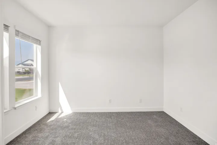 White painted walls with gray carpet flooring, featuring a large window with blinds and white trim