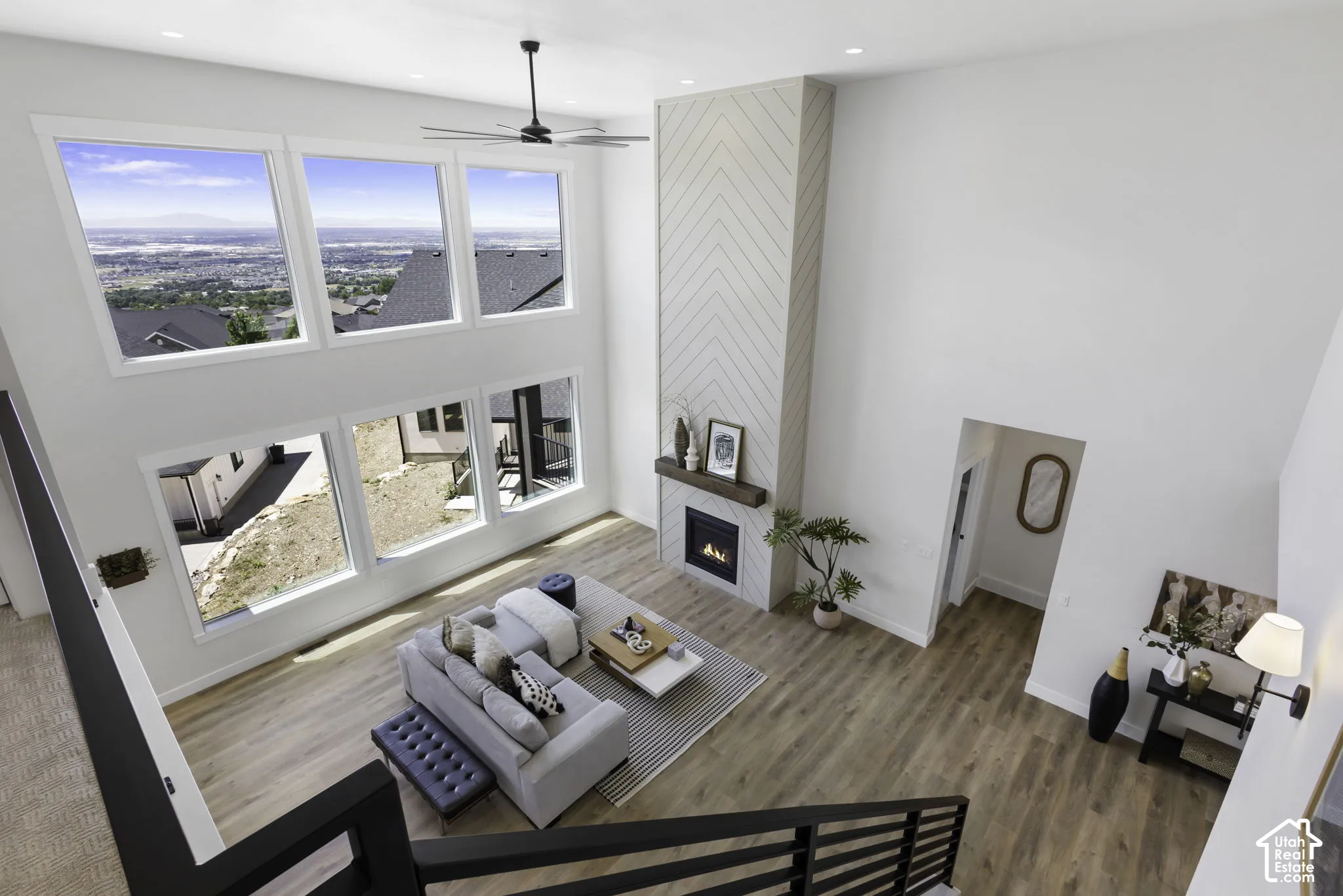 Soaring two-story living space with expansive multi-pane windows showcasing expansive landscape vistas, warm wood-finish flooring throughout, and a gas fireplace with stone surround