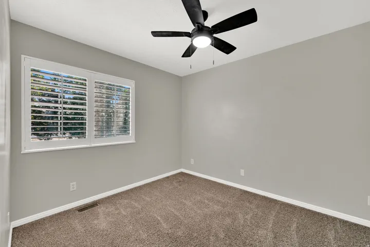 Window with white plantation shutters, neutral tone walls, ceiling-mounted fan with integrated lighting, and carpeted flooring