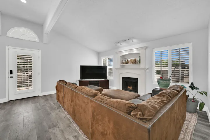 Vaulted ceiling with exposed beams, wood-finish flooring, white fireplace with mantle and built-in alcove, window with white plantation shutters, and a white paneled door with an arched transom