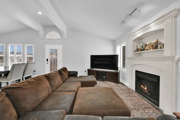 Vaulted ceiling with exposed beams, decorative fireplace with mantel and built-in shelving, white plantation shutters, and multiple windows