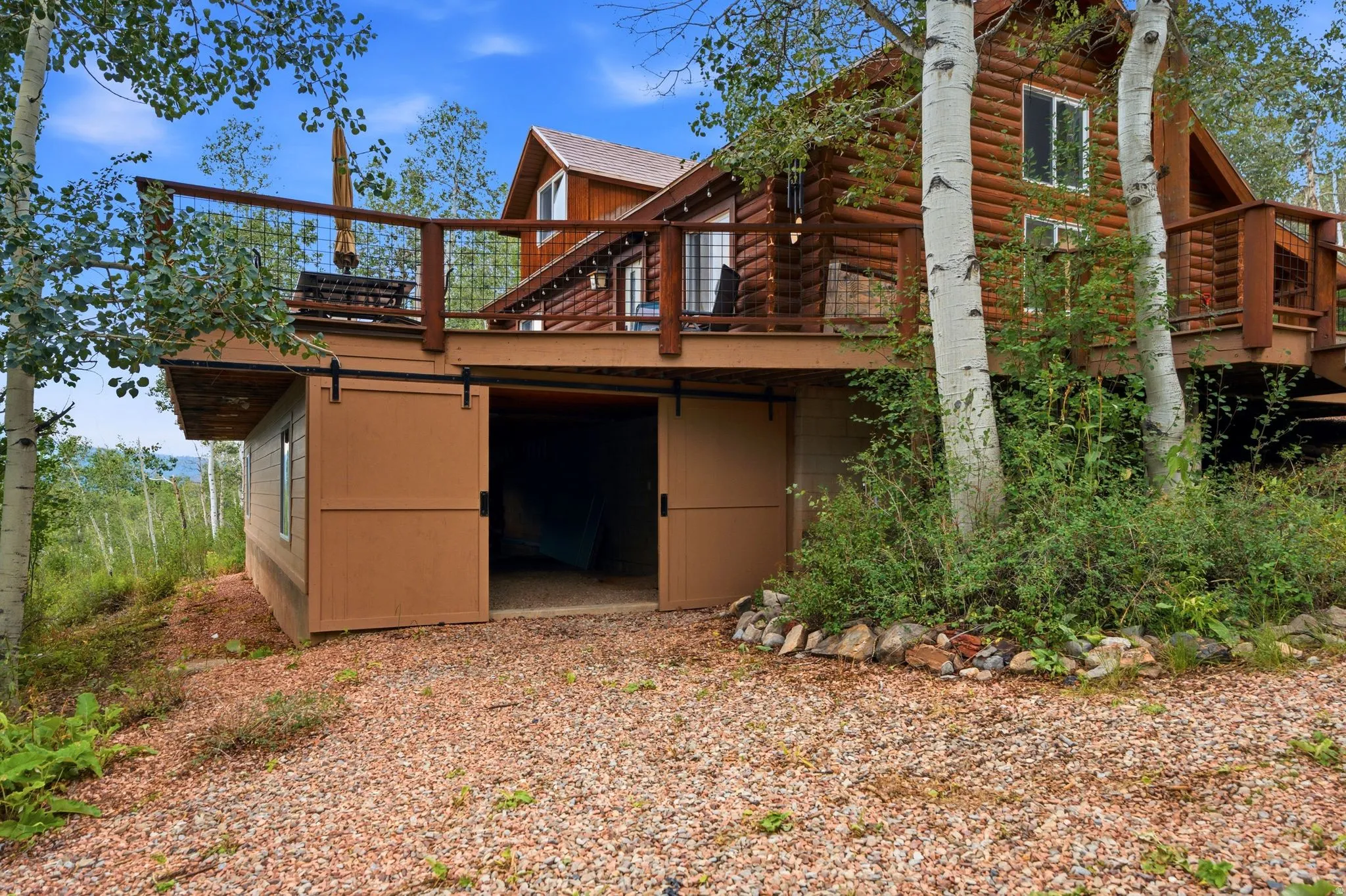 Log cabin exterior with an elevated wood deck featuring a railing, ground-level storage with barn-style sliding doors, and surrounding mature trees