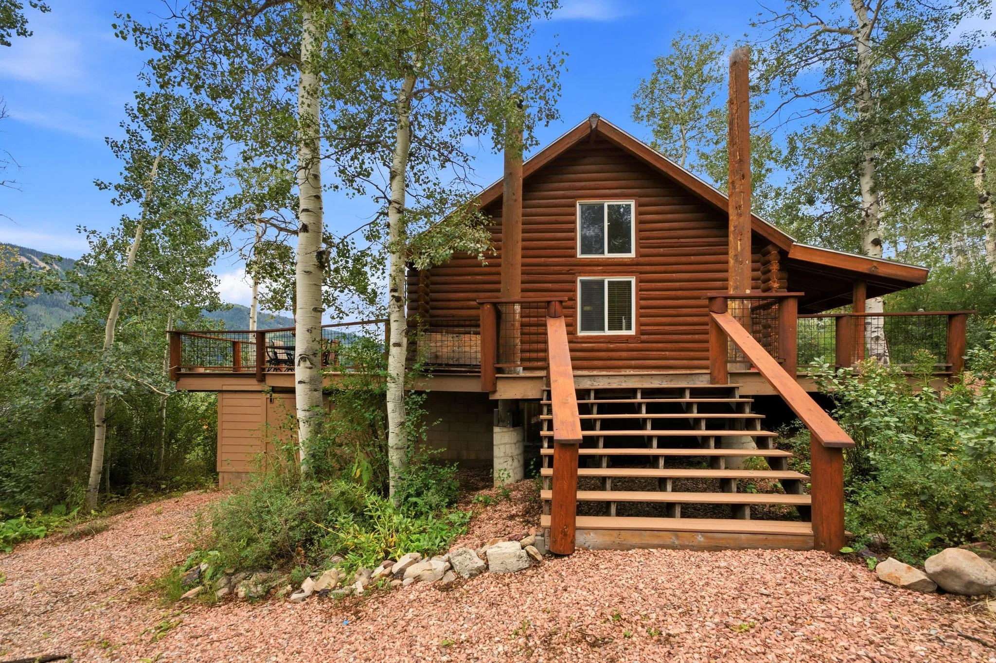 Wood log cabin exterior with a raised deck, outdoor stairs, and surrounding mature trees