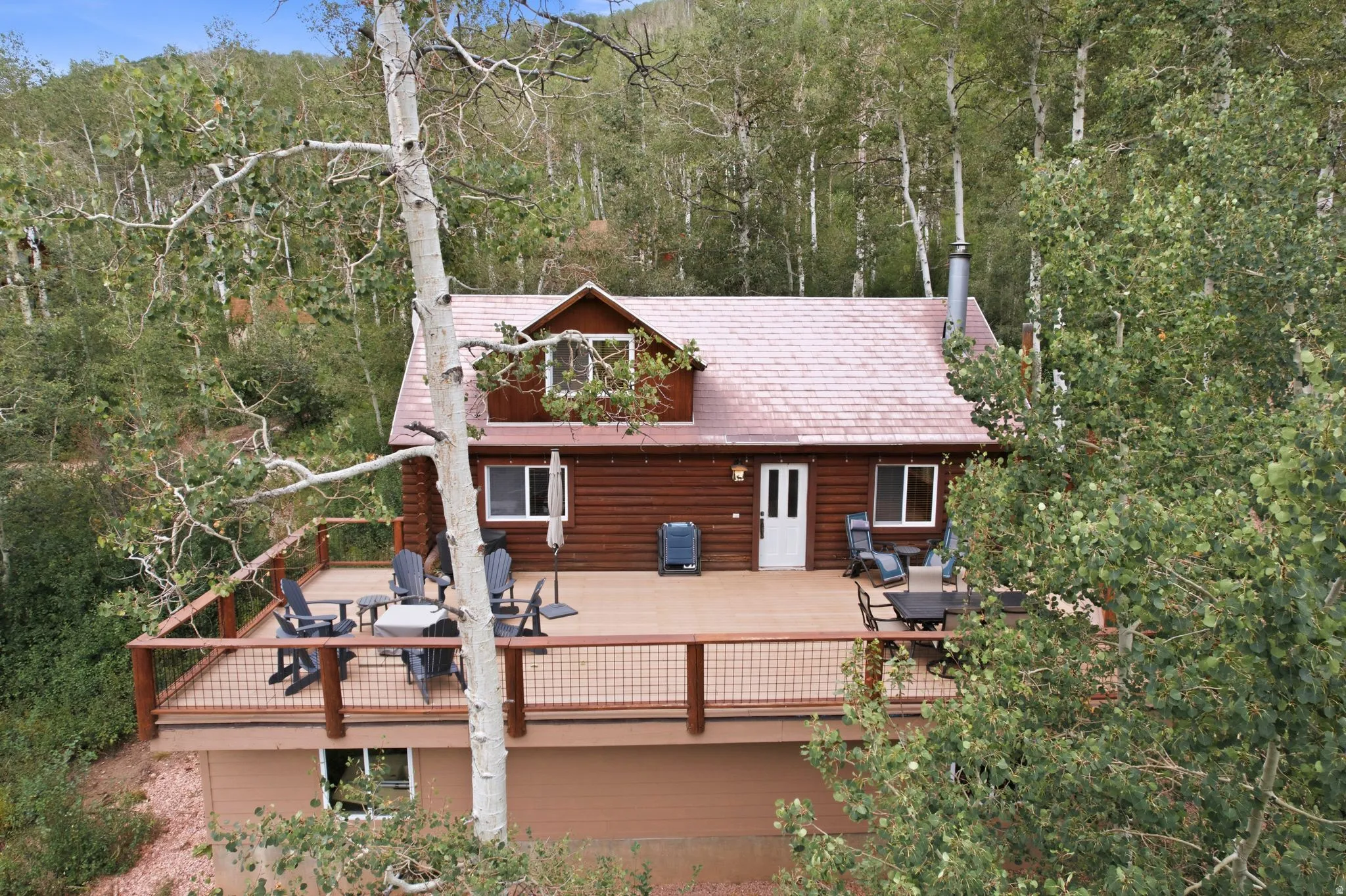 Log-sided exterior with a white entry door, large front deck with dark wood railing and metal infill, dormer window, and a light-colored shingle roof