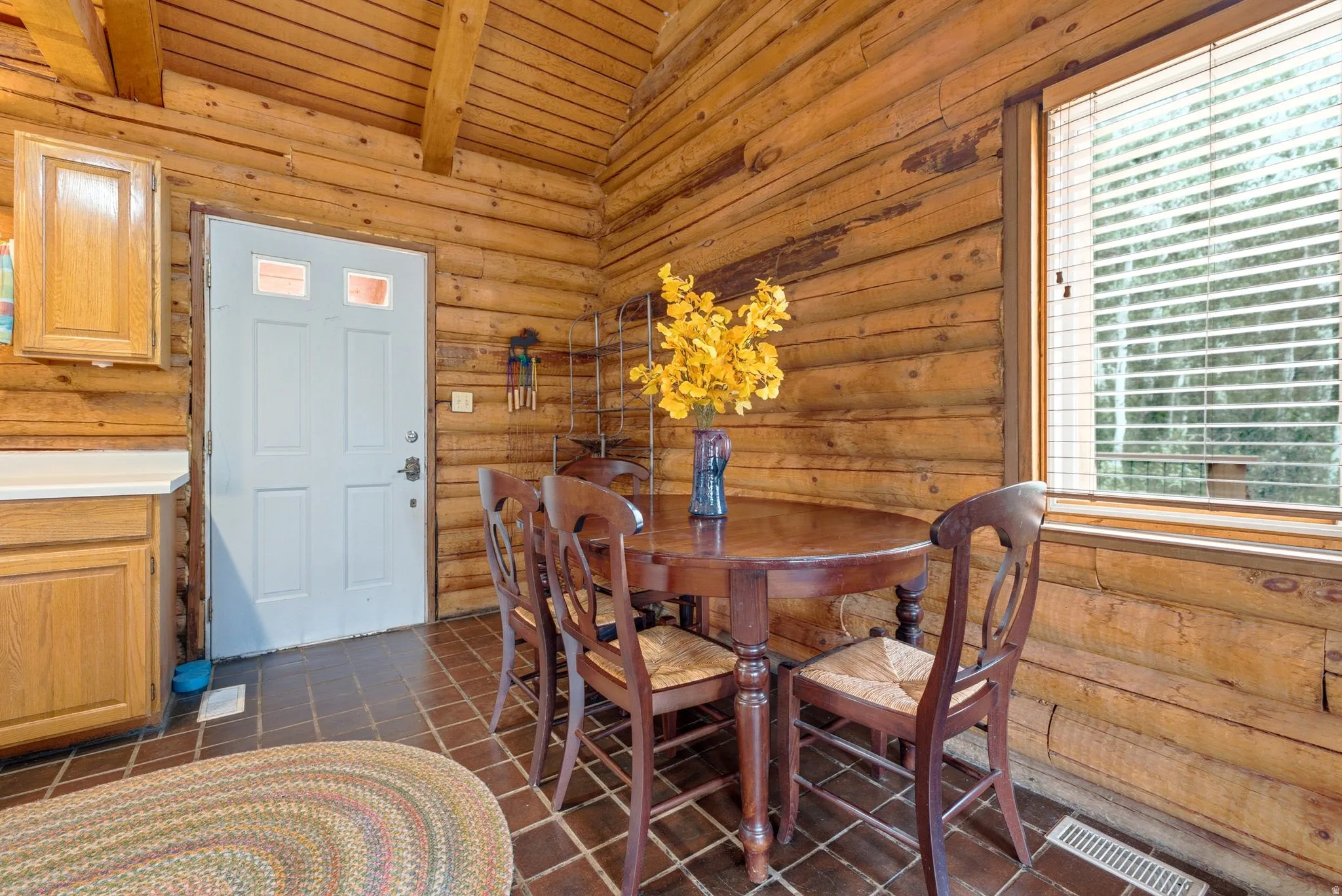 Log wall construction with exposed ceiling beams, a window with horizontal blinds, and a light-colored entry door