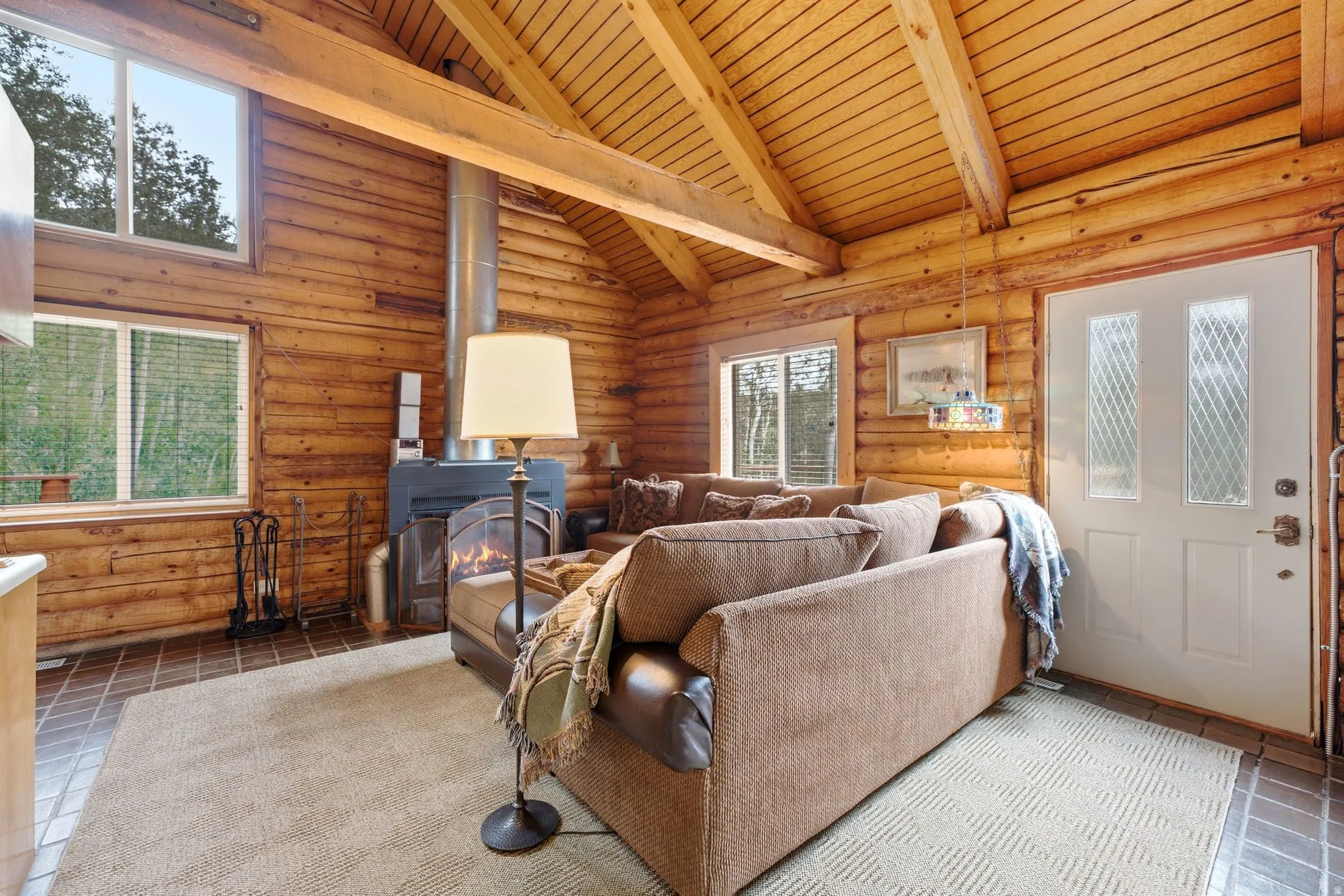 Log wall construction, exposed wood beam ceiling, wood burning stove, multiple windows, and a white entry door with decorative glass inserts
