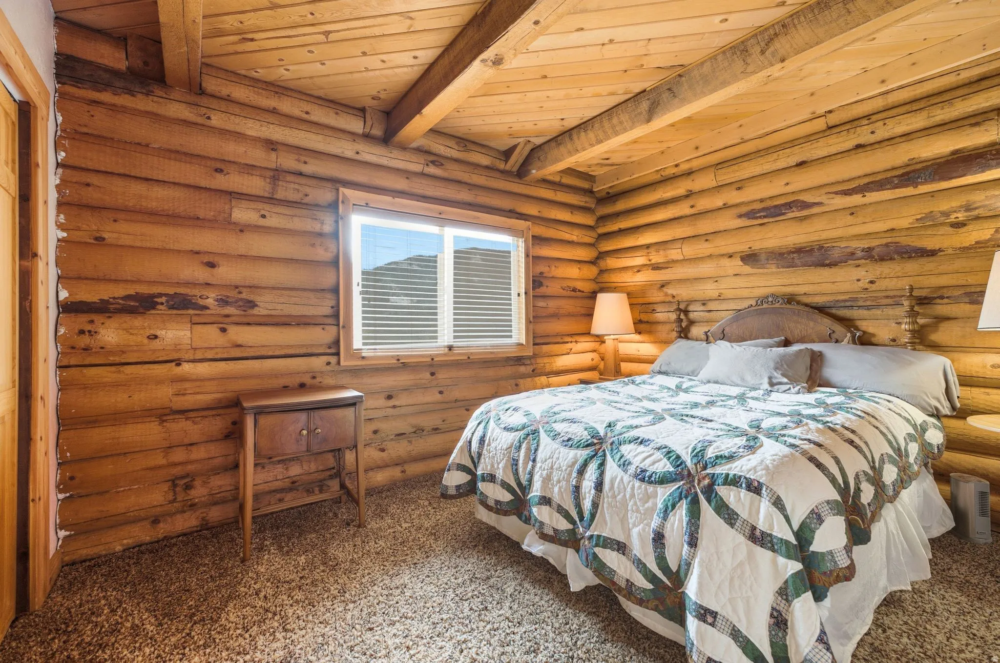 Rustic log cabin bedroom featuring hand-hewn timber walls and exposed beam ceiling, carpeted flooring, casement window with operable shutters, and built-in bedside table with integrated lamp fixture