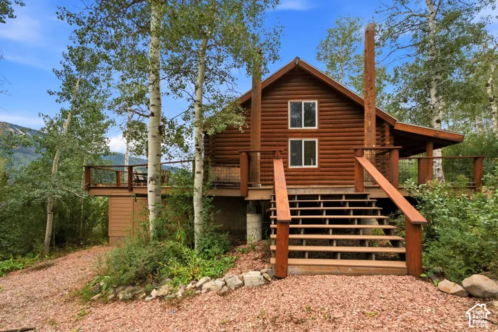 Wood log cabin exterior with a raised deck, outdoor stairs, and surrounding mature trees